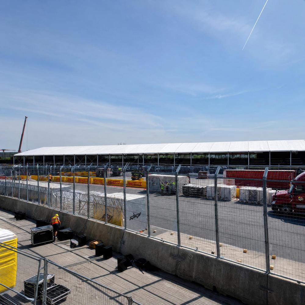 A ground-level, side-view shot of a race track paddock area. A large, multi-story temporary structure with glass walls runs along the track, separated by a metal fence.